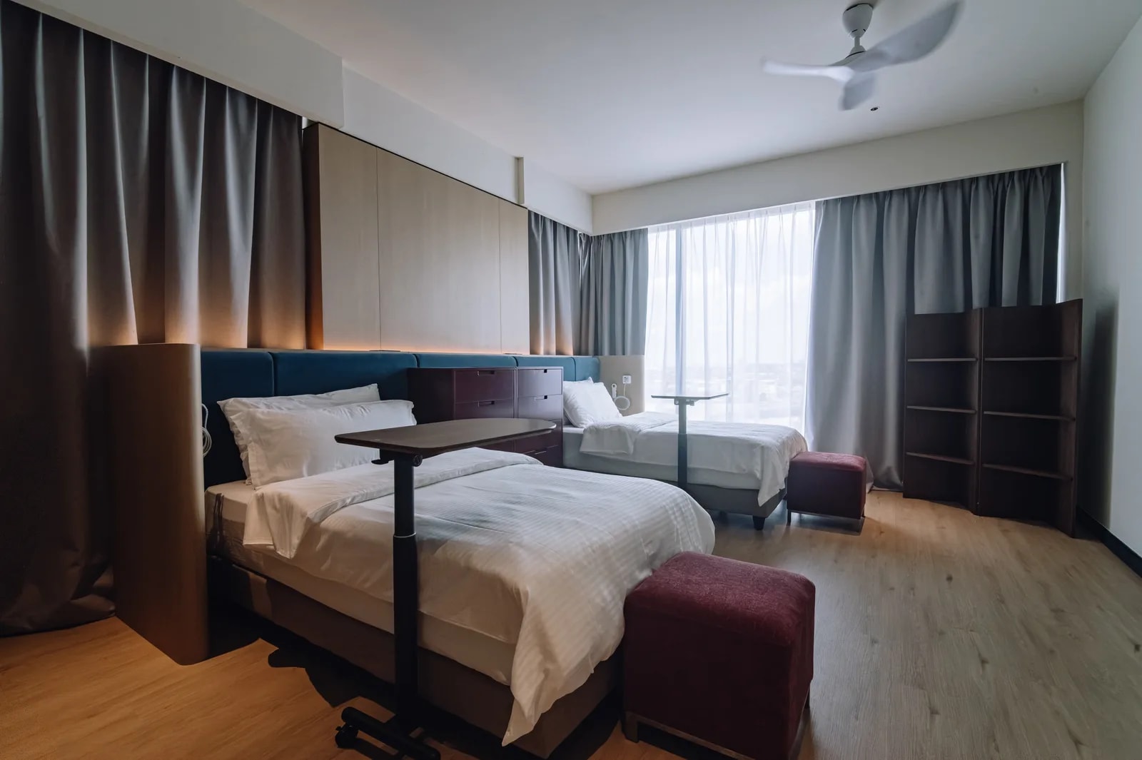 Twin room at Care Collective with two beds, overbed tables, and a window over Kuching