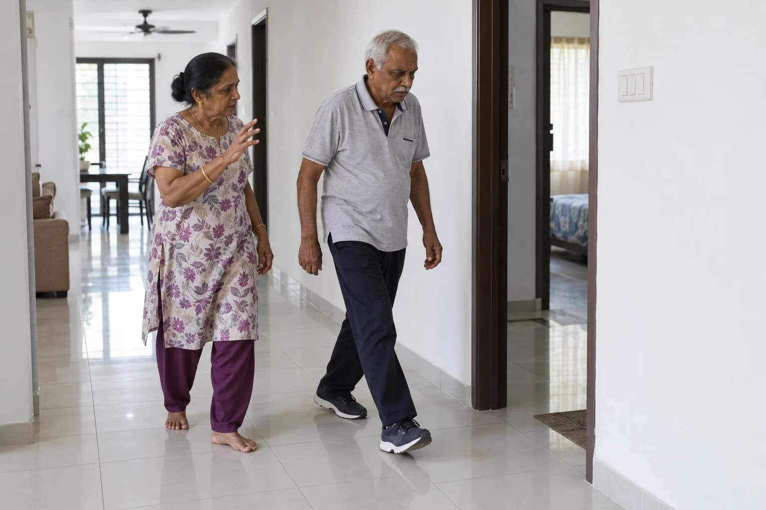 In a hallway at home, an older man walks with long steps while his wife walks beside him and gestures to guide him