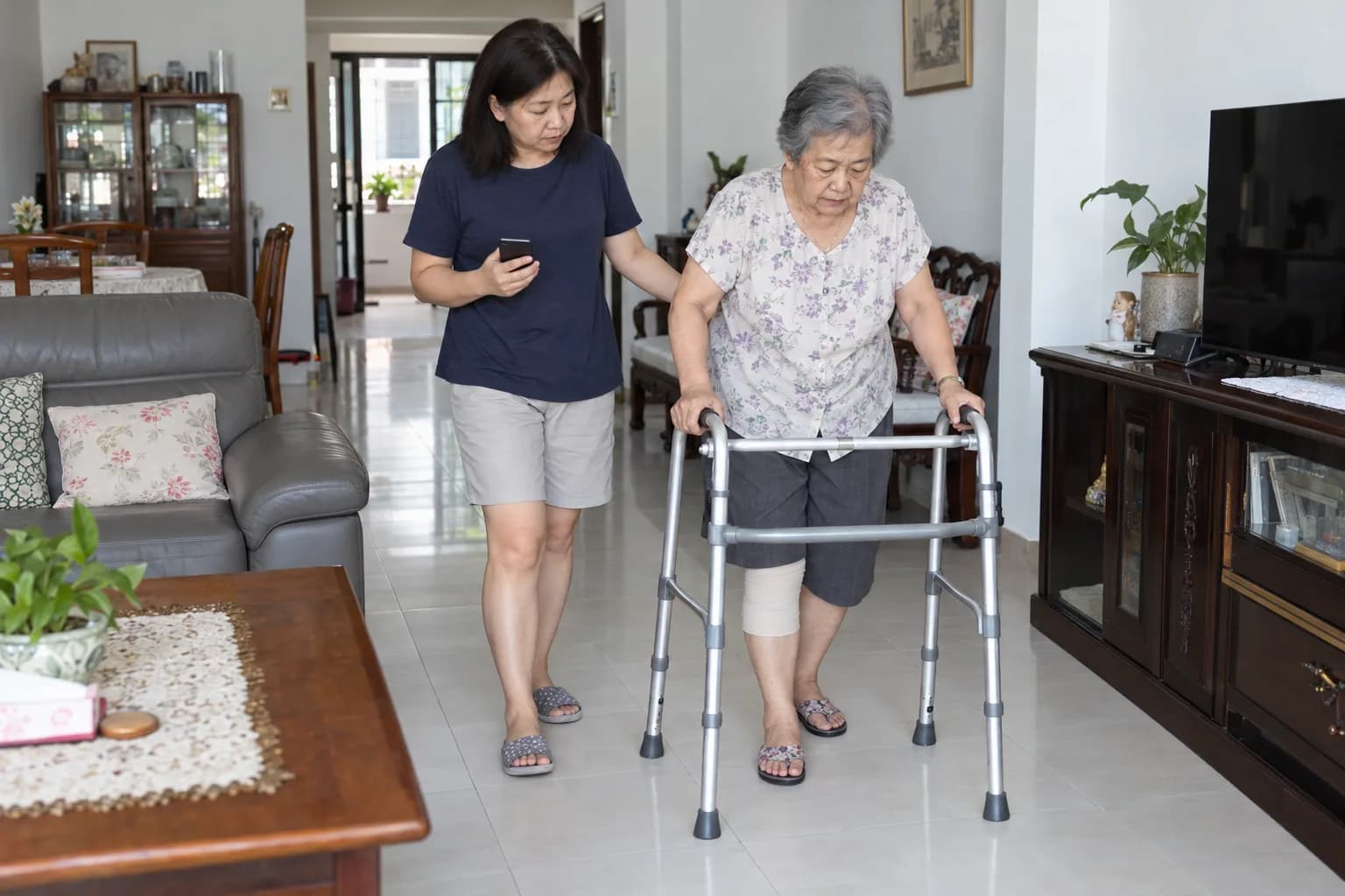 In a living room, an older woman with a bandaged knee walks with a walking frame while her daughter walks beside her