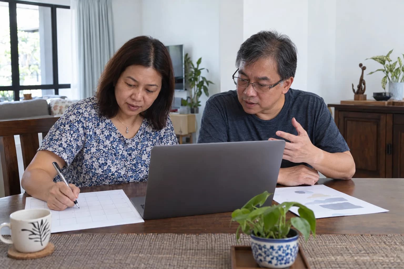 A couple at a dining table plan the weeks ahead, one writing on a calendar sheet while the other talks over a laptop