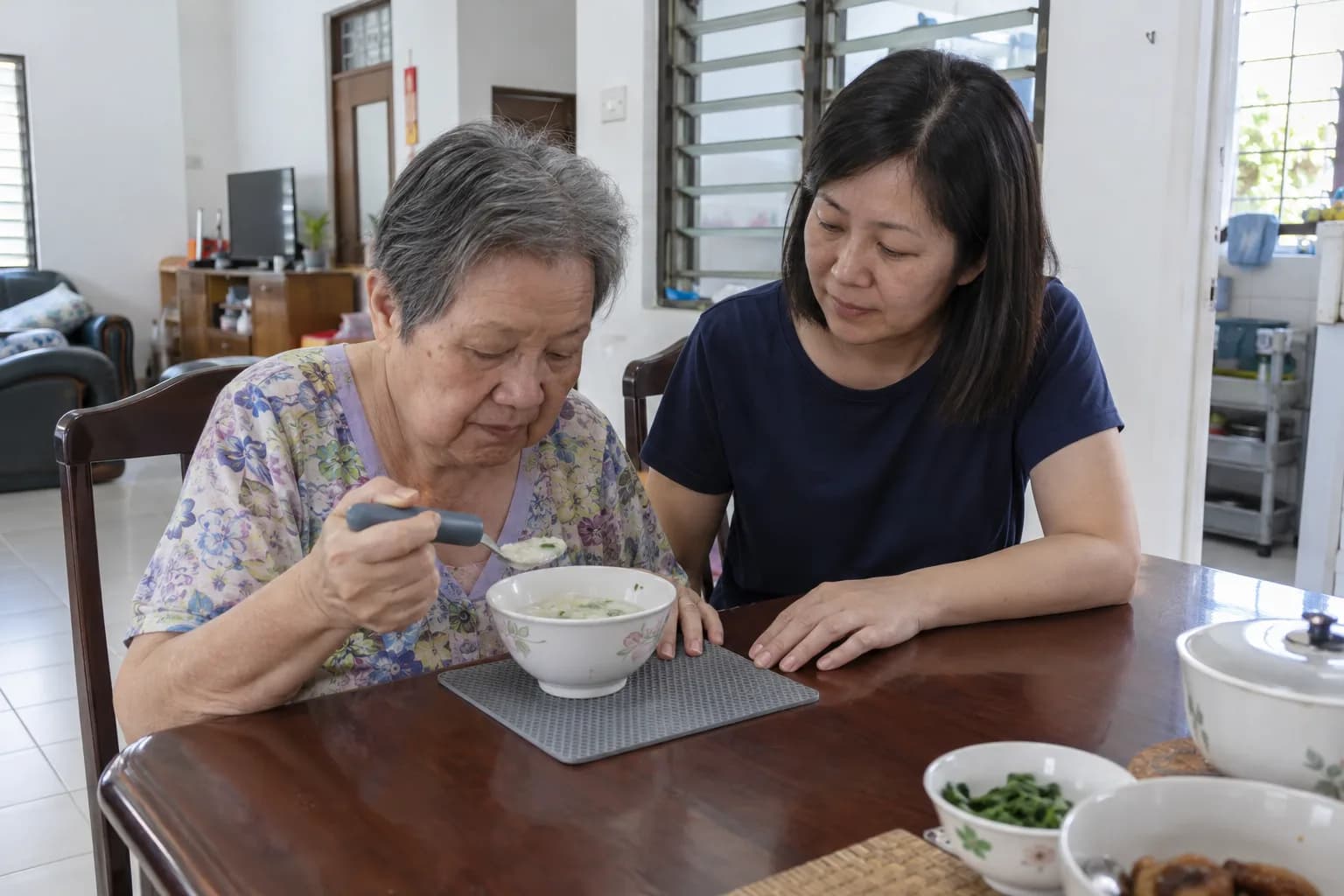 At a dining table at home, an older woman eats soup with a thick-handled spoon while her daughter sits beside her