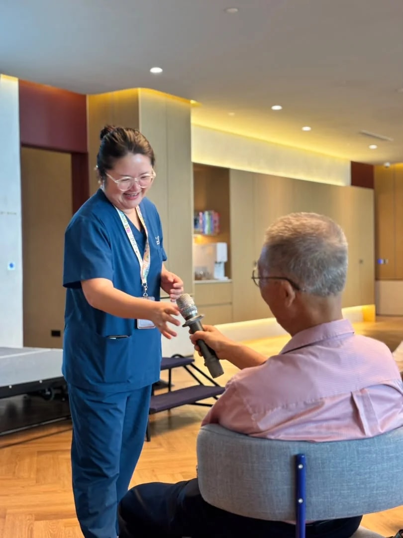 A caregiver in blue scrubs smiles as she hands a microphone to a seated member, seen from behind, in the Clubhouse lounge