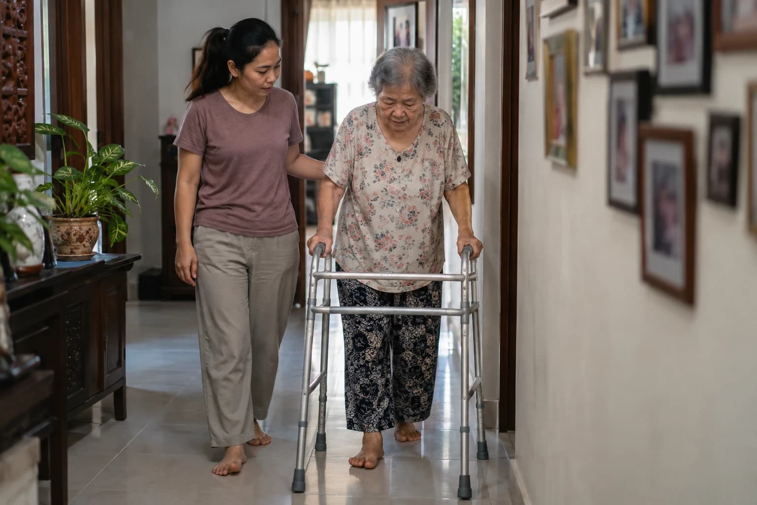A young helper walks slowly beside an older woman using a walking frame along a corridor at home